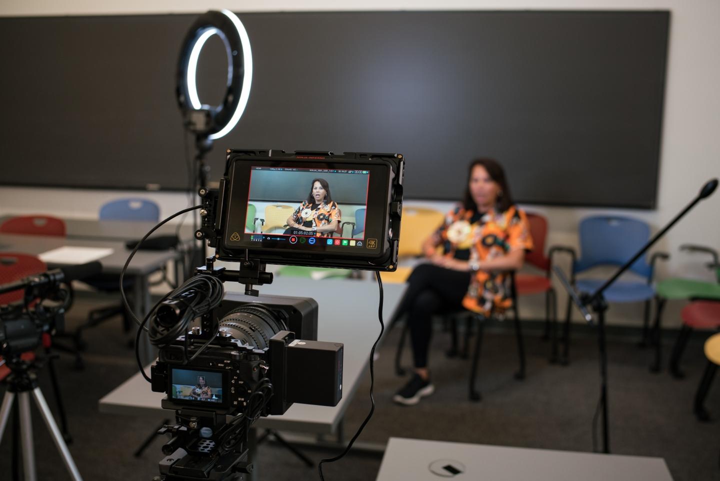 Alumna sitting in chair in classroom being videotaped for interview.